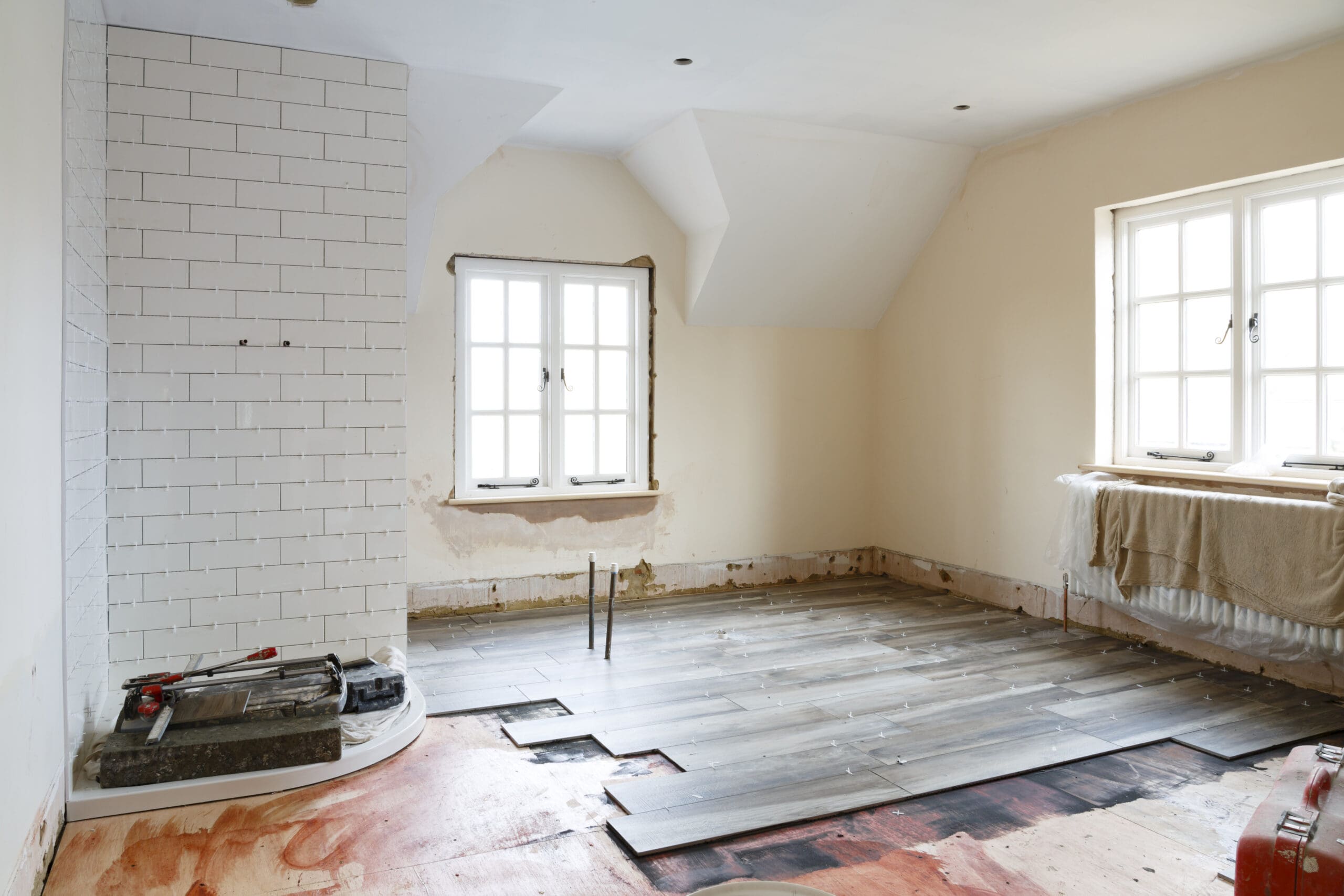 A room under renovation with bare walls and two windows. Partially installed wooden flooring is visible, and tiles are being laid against one wall. Construction tools are scattered across the floor, and a window on the right has a towel draped over it, reflecting ongoing refurbishments.