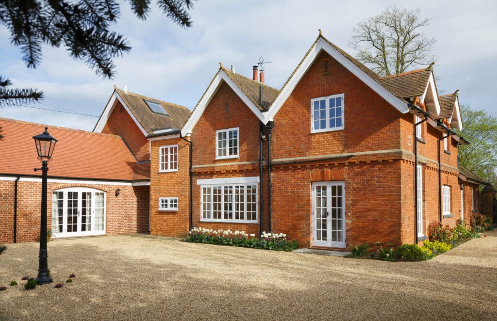 A large red-brick house with white-framed windows and doors, steep gabled roofs, and a gravel driveway showcases quality building services; flowerbeds with greenery line the home, while bright skies shine above a black lamppost.