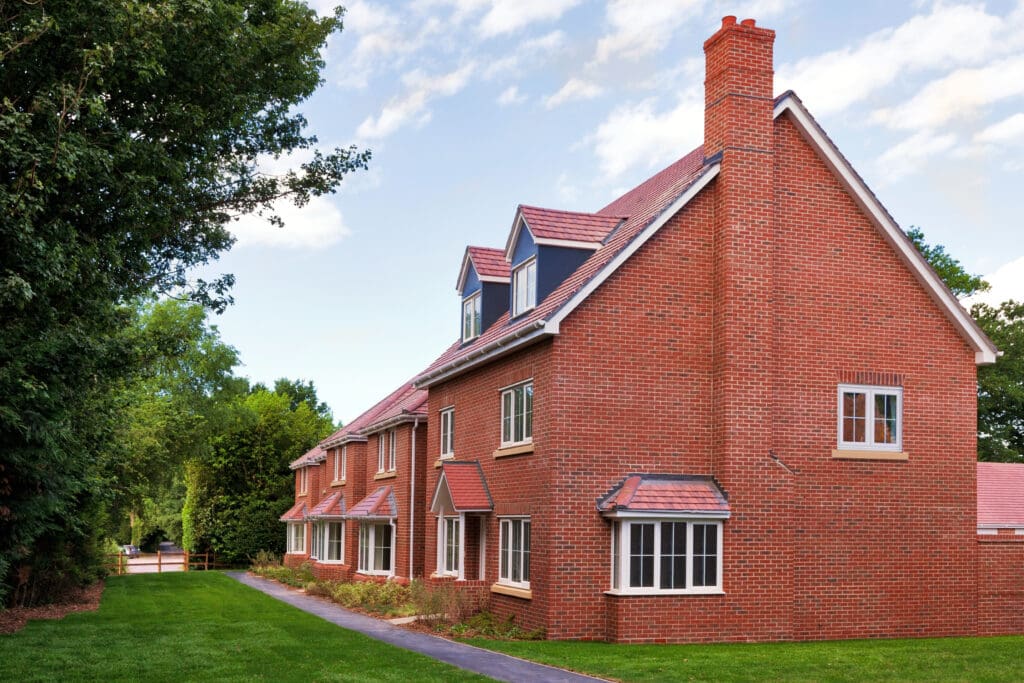 A modern two-story red-brick house with multiple gabled roofs and white-trimmed windows, surrounded by green lawn and trees under a partly cloudy sky.