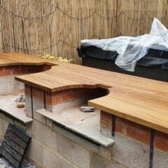A person stands next to an outdoor kitchen under construction, featuring wooden countertops atop brick and block bases, with tools and materials scattered nearby and a bamboo privacy screen in the background.