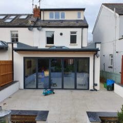 A modern house extension with large sliding glass doors opens onto a spacious patio. The patio is tiled, with a blue children's scooter and a bin visible. Neighboring houses and wooden fences flank the area.