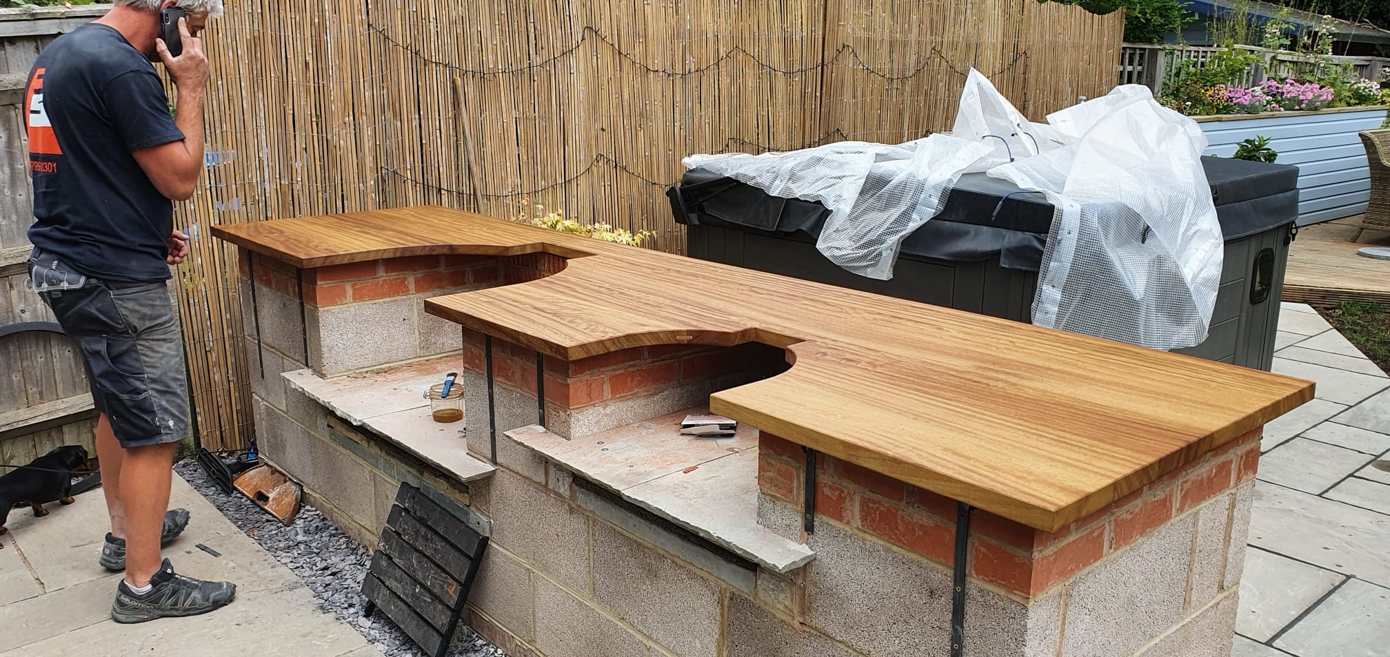 A person stands next to an outdoor kitchen under construction, featuring wooden countertops atop brick and block bases, with tools and materials scattered nearby and a bamboo privacy screen in the background.