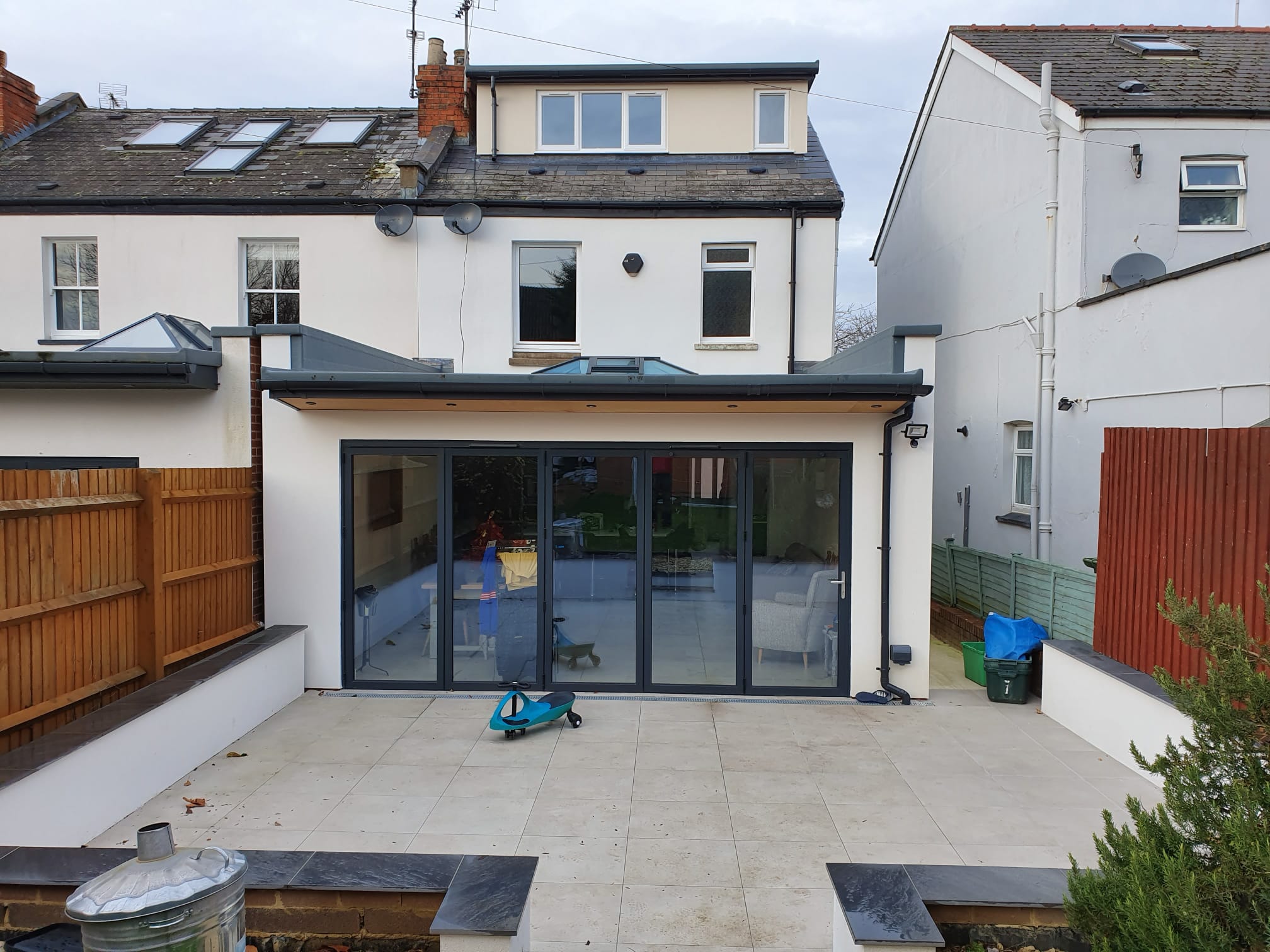 A modern house extension with large sliding glass doors opens onto a spacious patio. The patio is tiled, with a blue children's scooter and a bin visible. Neighboring houses and wooden fences flank the area.