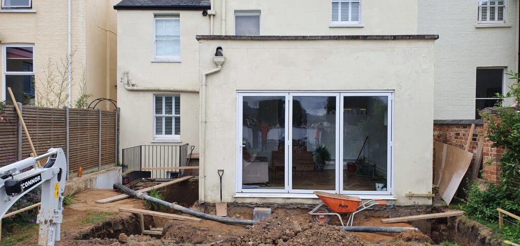 A house with large glass doors and an extension under construction. The ground is dug up, exposing soil and pipes, with a wheelbarrow in front. Construction materials are scattered around the site.