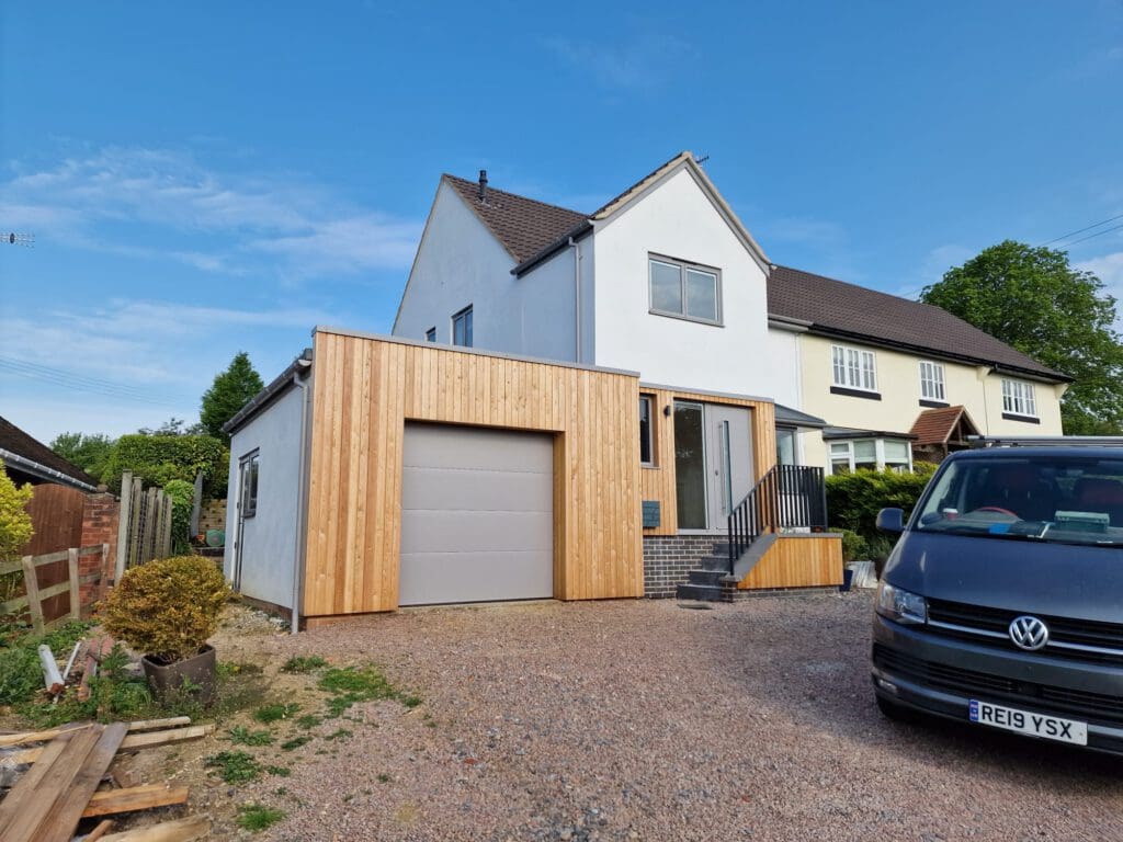 A modern house with a white upper facade and a new wooden extension featuring a garage door. A black Volkswagen van is parked in the gravel driveway under a clear blue sky.