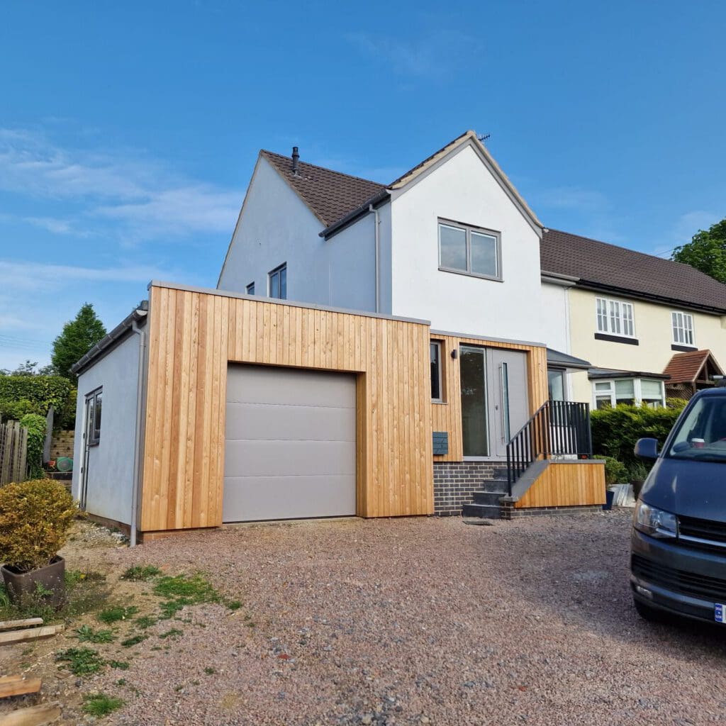 A modern house with a white upper facade and a new wooden extension featuring a garage door. A black Volkswagen van is parked in the gravel driveway under a clear blue sky.