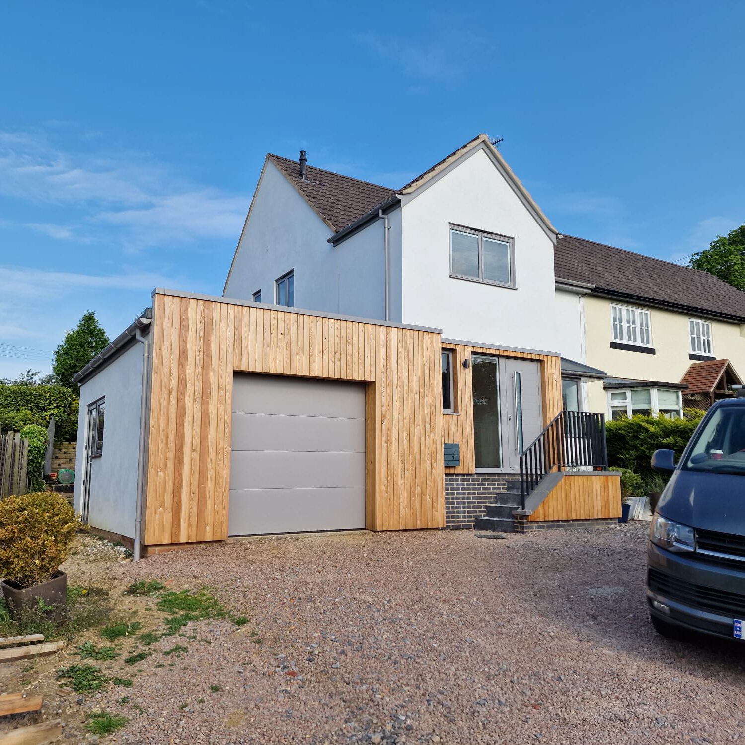 A modern house with a white upper facade and a new wooden extension featuring a garage door. A black Volkswagen van is parked in the gravel driveway under a clear blue sky.