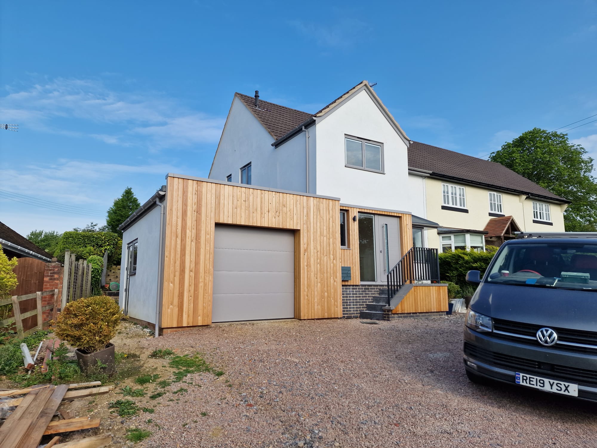 A modern house with a white upper facade and a new wooden extension featuring a garage door. A black Volkswagen van is parked in the gravel driveway under a clear blue sky.