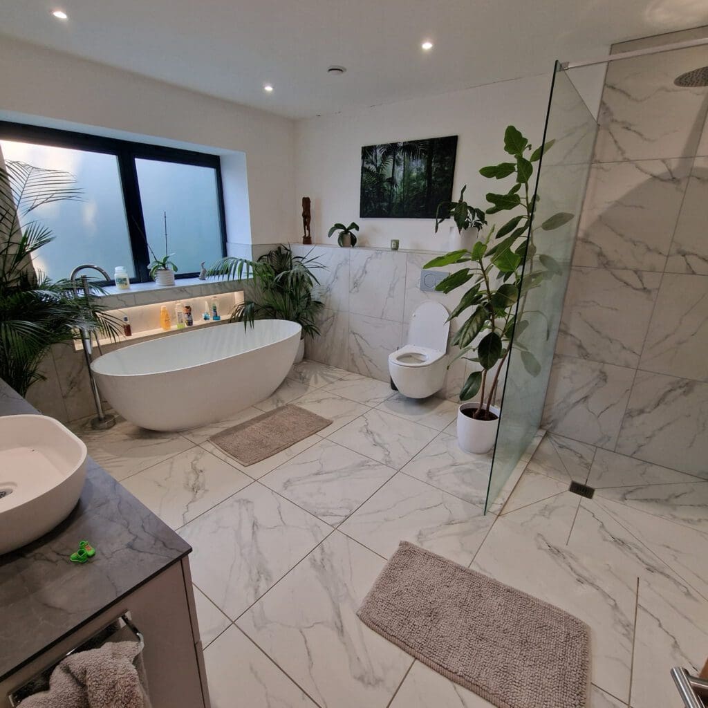 Modern bathroom with white marble tiles, a freestanding bathtub, glass shower, and toilet. Green plants and a large window add a natural touch. Soft bath mats are placed on the floor. A sink and countertop are visible.