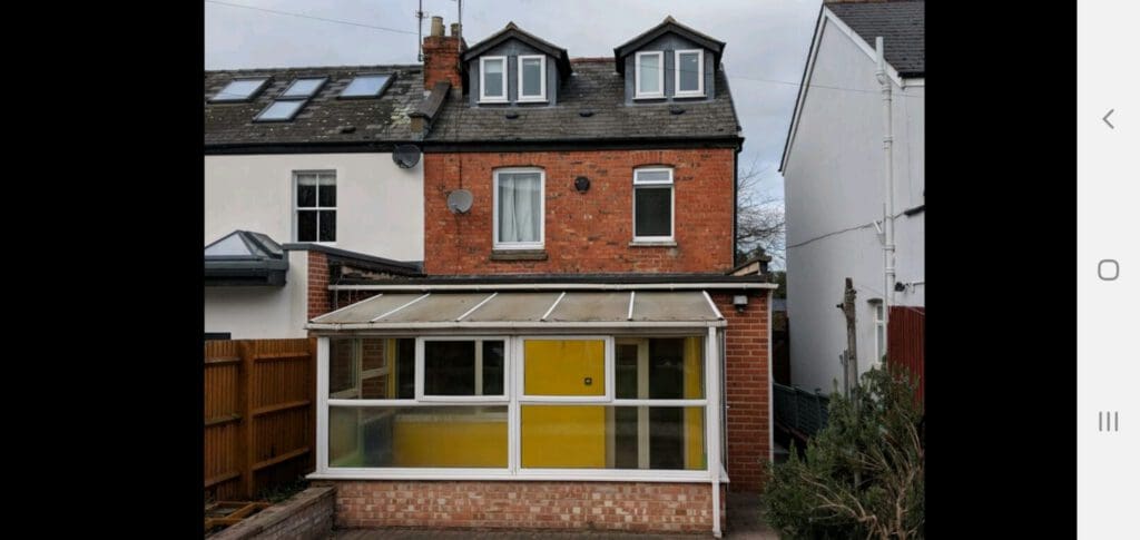 A two-story brick house with two dormer windows on the roof and a ground-floor sunroom extension featuring large yellow and white window panels. The house has a small paved area and a wooden fence on one side.