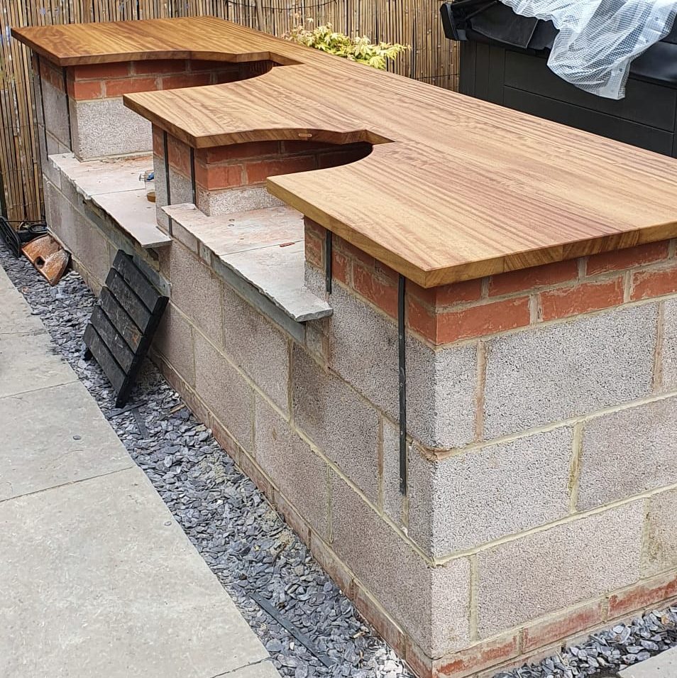 A partially completed outdoor kitchen with a brick and concrete block base, wooden countertops, and a bamboo privacy fence in the background, surrounded by greenery.