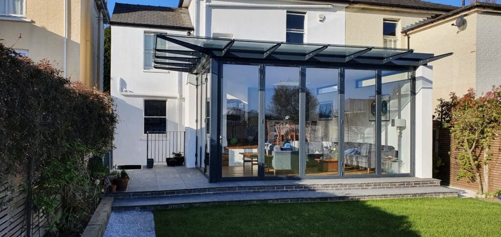 A modern glass extension with a sloped roof at the back of a white house, opening onto a garden with a lawn; the interior has sofas and a table, visible through large glass doors and windows.