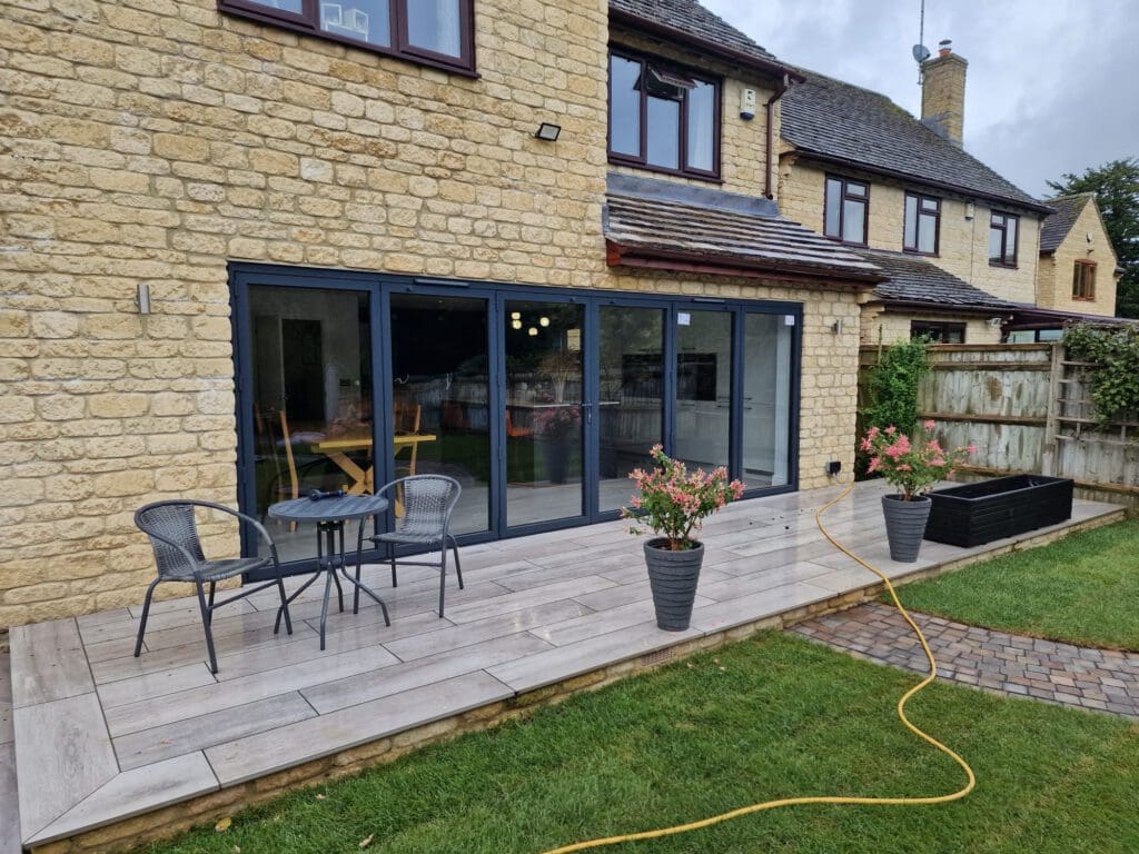 A stone house with large glass doors opens onto a wooden deck. The deck has two chairs, a small table, potted plants, and a yellow garden hose. A flowerbed runs along a wooden fence in the grassy yard.