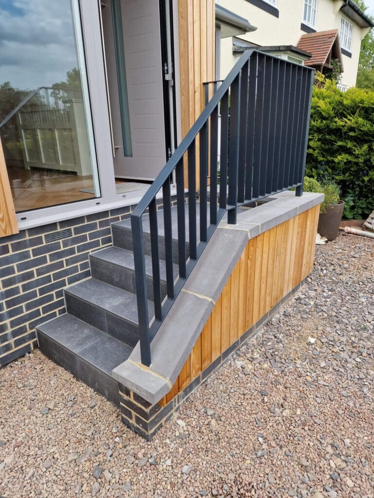 A small outdoor staircase with dark railings and gray tiles leads to a door on a modern house with wood and brick siding, set above a gravel ground.