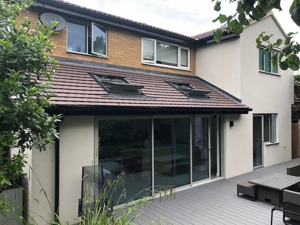 A modern two-story house with cream walls, brick accents, and brown roof tiles features large sliding glass doors and skylights, opening onto a gray deck with outdoor seating and surrounded by greenery.