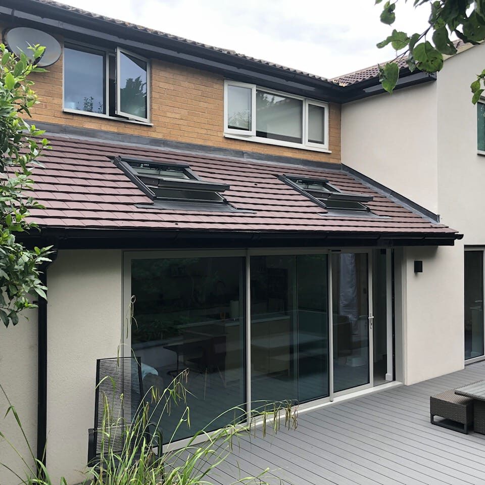 A modern two-story house with cream walls, brick accents, and brown roof tiles features large sliding glass doors and skylights, opening onto a gray deck with outdoor seating and surrounded by greenery.