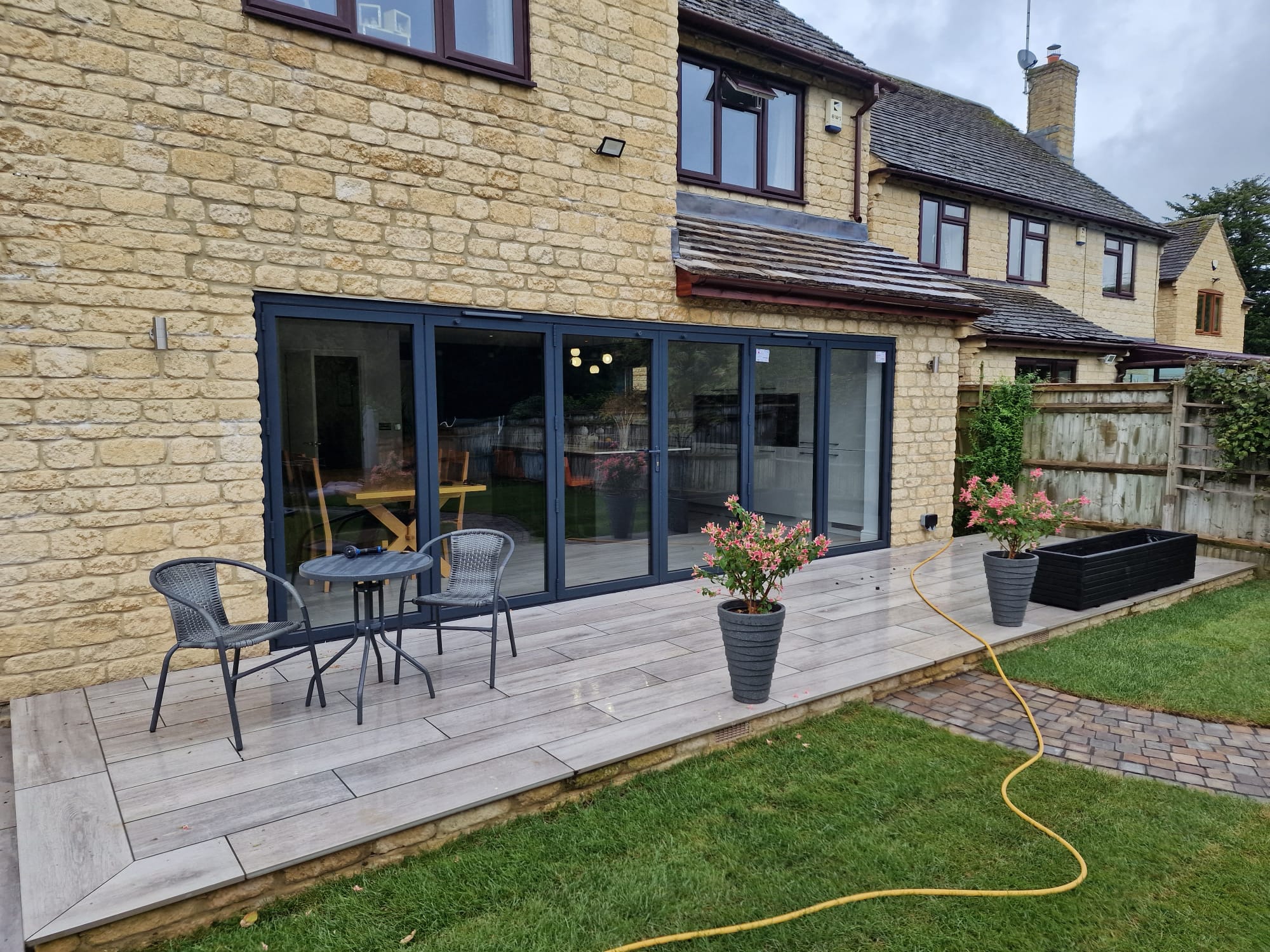 A stone house with large glass doors opens onto a wooden deck. The deck has two chairs, a small table, potted plants, and a yellow garden hose. A flowerbed runs along a wooden fence in the grassy yard.
