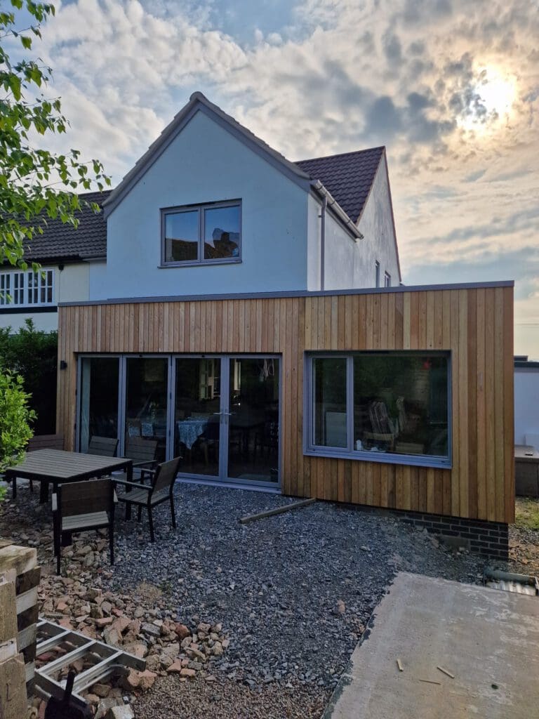 A modern house extension with vertical wooden cladding, large glass sliding doors, and a gravel patio area with outdoor furniture. The sun is partially visible through a cloudy sky above the house.