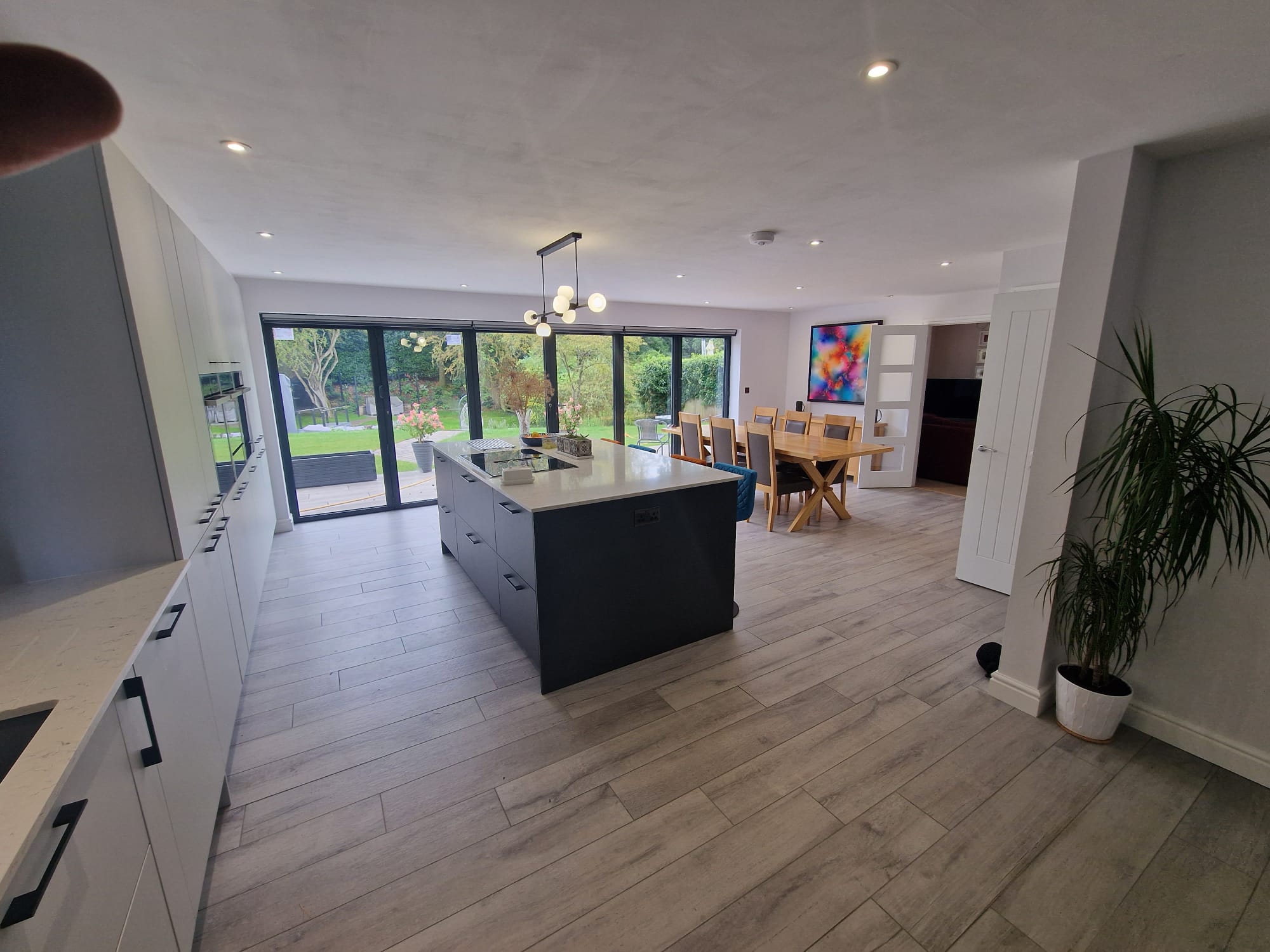 Modern open-plan kitchen and dining area with light wood flooring, a large island, sleek white cabinets, sliding glass doors to a garden, a wooden dining table, colorful artwork, and a tall potted plant.