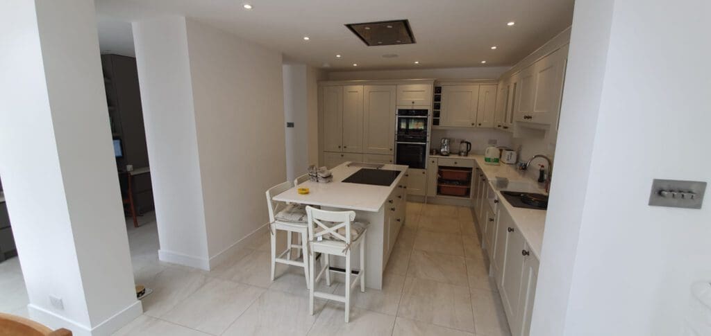 Modern white kitchen with an island, two chairs, built-in oven, stove, sink, kettle, and toaster. The room is bright with recessed lighting and has light-colored tile flooring. A door opens to another room on the left.