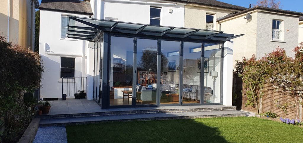A modern glass conservatory with a slanted glass roof is attached to the back of a white house, opening onto a stone patio and a green lawn. The interior shows sofas, a table, and a sunlit, cozy setting.