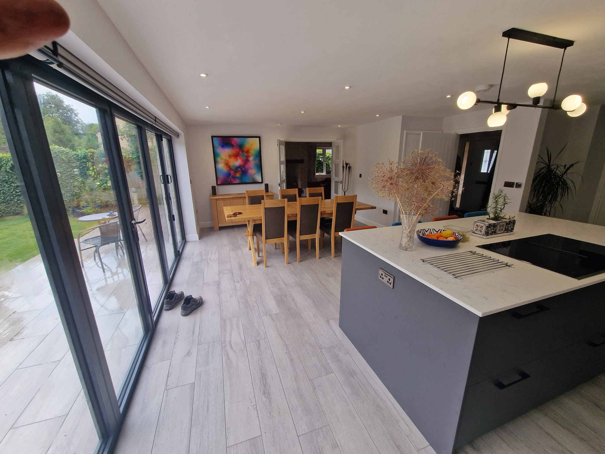 Modern open-plan kitchen and dining area with light wood floors, a large island, wooden dining table, colorful wall art, large glass doors to a garden, and shoes by the entrance.