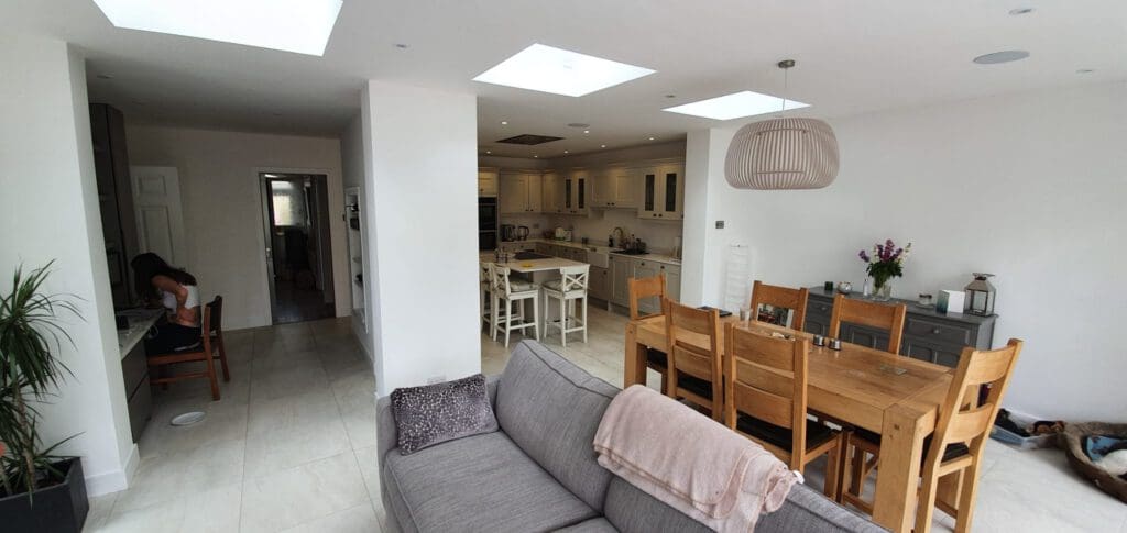 Modern open-plan kitchen and dining area with a wooden table, chairs, grey sofa, and someone working at a desk in the corner. The room is bright with white walls, ceiling lights, and skylights.