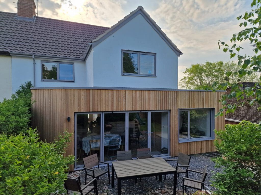 A modern house with a white upper story and wooden paneling on the lower level, large sliding glass doors, and an outdoor patio area with tables and chairs surrounded by greenery under a partly cloudy sky.