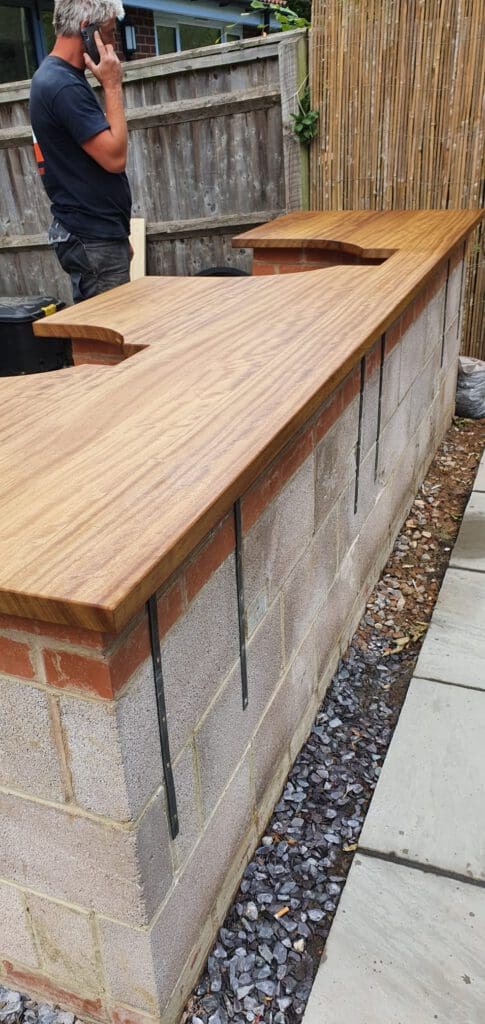 A man stands beside an outdoor bar counter made of concrete blocks with a smooth wooden top and metal supports, set against a wooden fence and bamboo privacy screen.