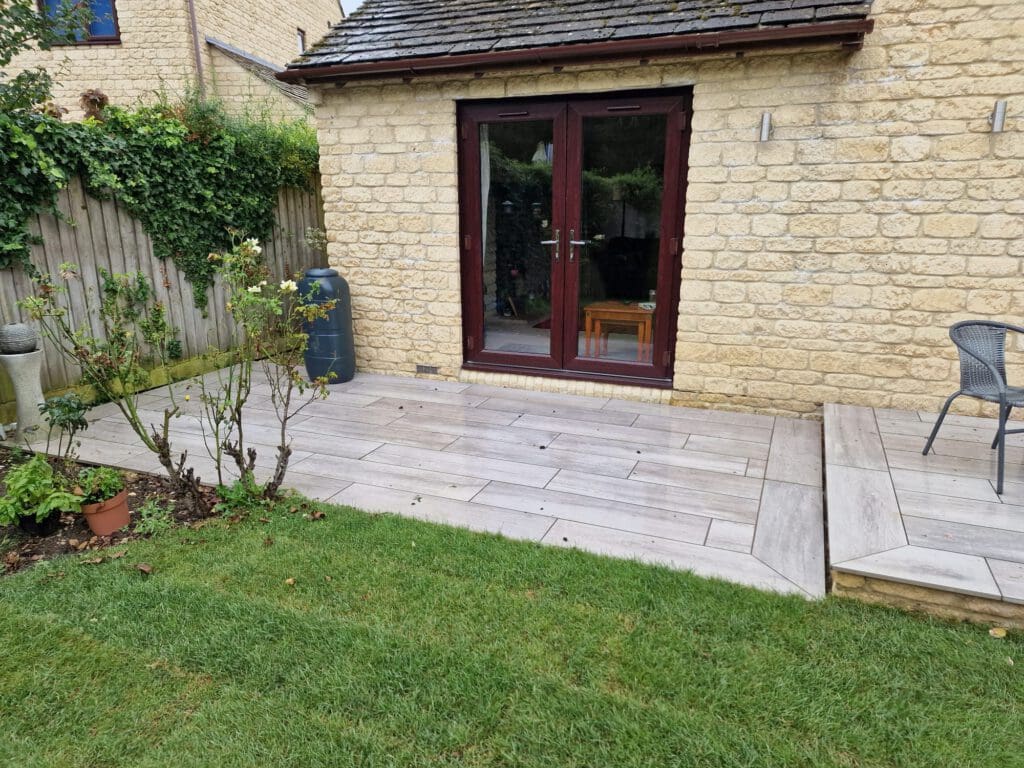 A stone patio with light-colored tiles borders a garden with grass, rose bushes, and a wooden fence. There's a small round table and chair, and double glass doors lead into a stone building.