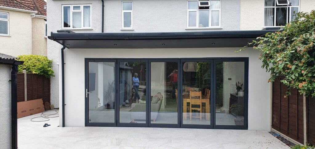 A modern house extension with large glass folding doors, reflecting people and a garden outside. Inside, a wooden dining table and chairs are visible. The exterior features light walls and a tiled patio.