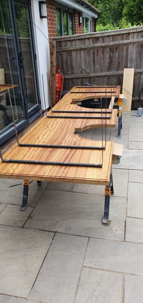 A large wooden board rests on workbenches outdoors, with metal rods forming rectangular frames attached to it. The setup is next to a wooden fence and a brick building with glass doors.