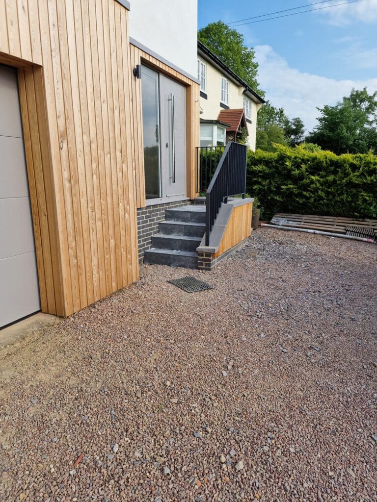 A set of gray stone steps with a black metal railing leads up to a glass door. The house has wood paneling and white walls, with gravel covering the ground and greenery in the background.