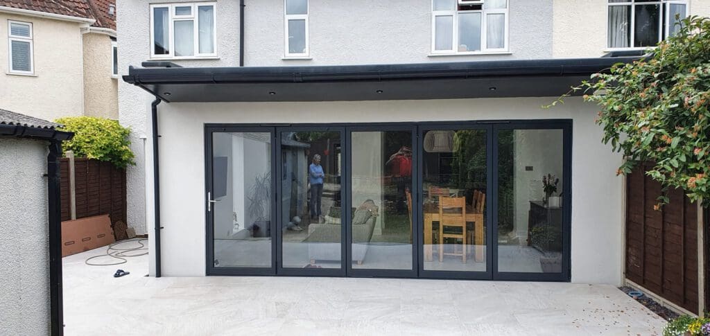A modern house extension with large glass folding doors opening onto a patio; inside, a man stands by a dining table with wooden chairs, and plants are visible on the right side.