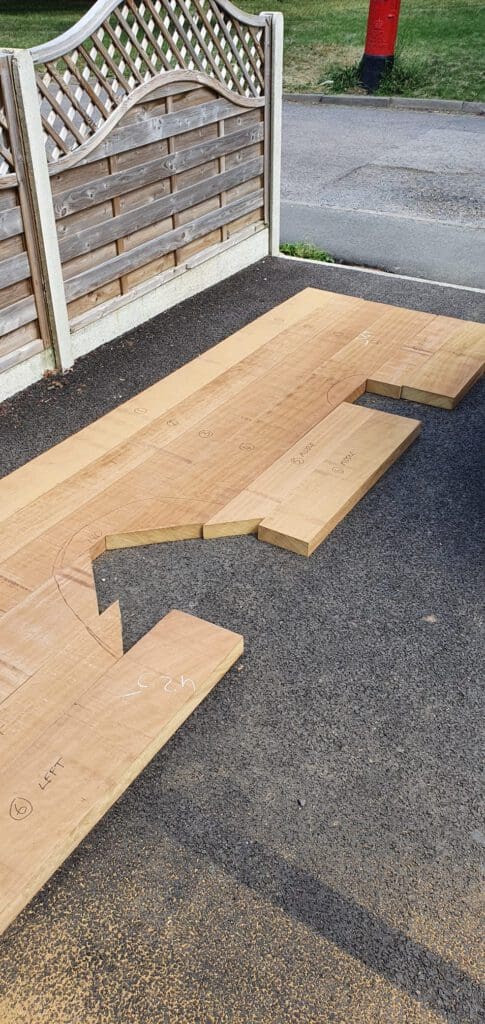 Several cut wooden boards are arranged on a driveway next to a wooden fence. The boards appear measured and labeled for a project, some with angled cuts and markings, possibly for construction or carpentry work.
