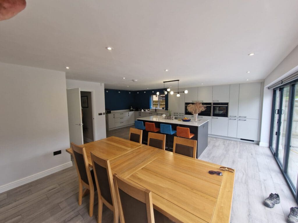 Spacious modern kitchen and dining area with a wooden table and six chairs, a kitchen island with orange and blue stools, white cabinets, and large sliding glass doors. Shoes and sunglasses are on the floor and table.