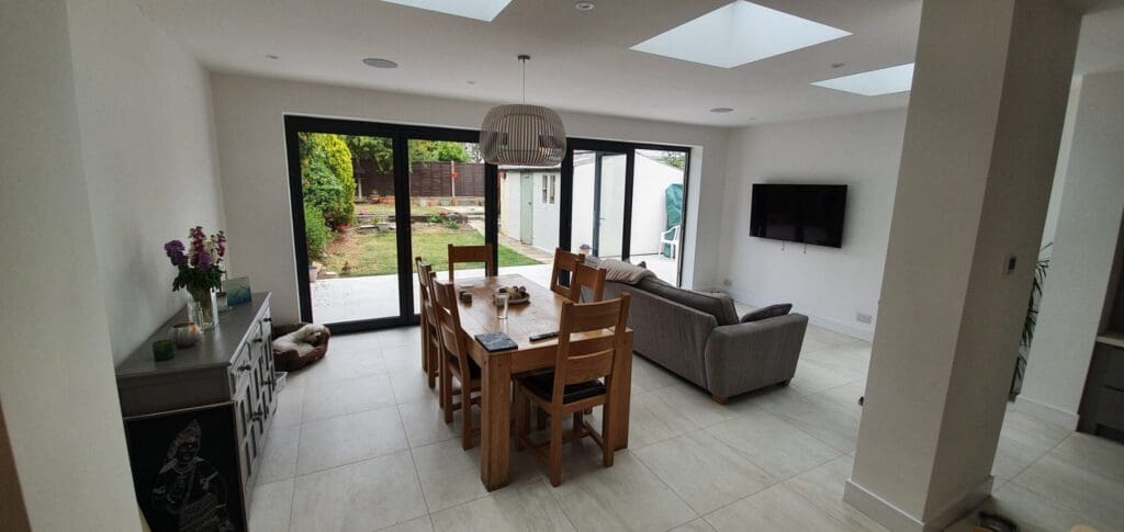 A modern, open-plan dining and living area with large glass doors to a garden. There’s a wooden dining table with chairs, a gray sofa facing a wall-mounted TV, and skylights above letting in natural light.