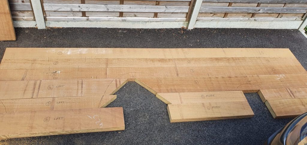 Large wooden planks with various measurements and markings in white chalk are laid out on a black asphalt surface near a wooden fence. Some planks have cut-out sections and labels like "LEFT" and "NOSE.
