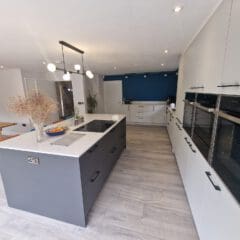 Modern kitchen with a large gray island, white countertops, built-in black ovens, light wood flooring, a blue accent wall, and a dining area with wooden table and chairs in the background. Bright, minimalistic design.