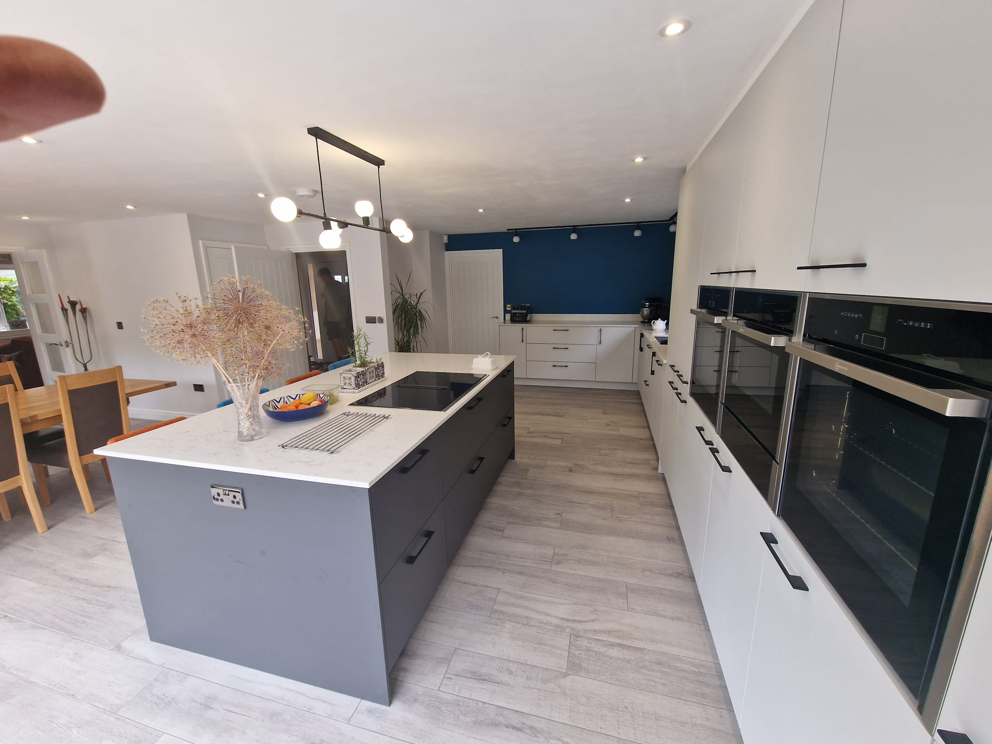 Modern kitchen with a large gray island, white countertops, built-in black ovens, light wood flooring, a blue accent wall, and a dining area with wooden table and chairs in the background. Bright, minimalistic design.