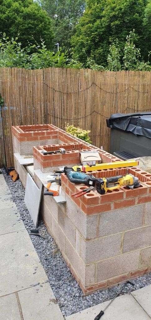 A partially constructed outdoor kitchen made of red and gray concrete blocks stands on gravel beside a bamboo privacy fence, with various tools including a level, drill, and saw resting on top.