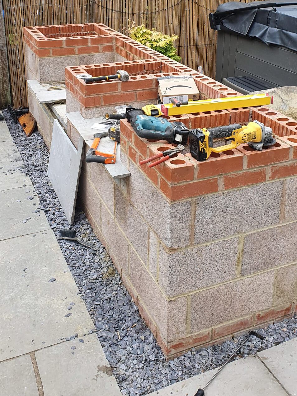 A partially constructed outdoor kitchen made of red and gray concrete blocks stands on gravel beside a bamboo privacy fence, with various tools including a level, drill, and saw resting on top.