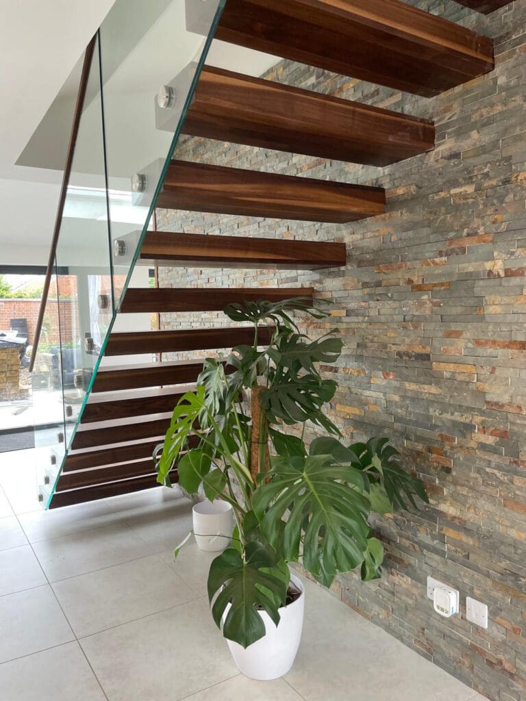 Modern floating wooden staircase with glass railing beside a stone-textured wall. Two large potted Monstera plants sit on the white tiled floor underneath the stairs. Natural light fills the space.
