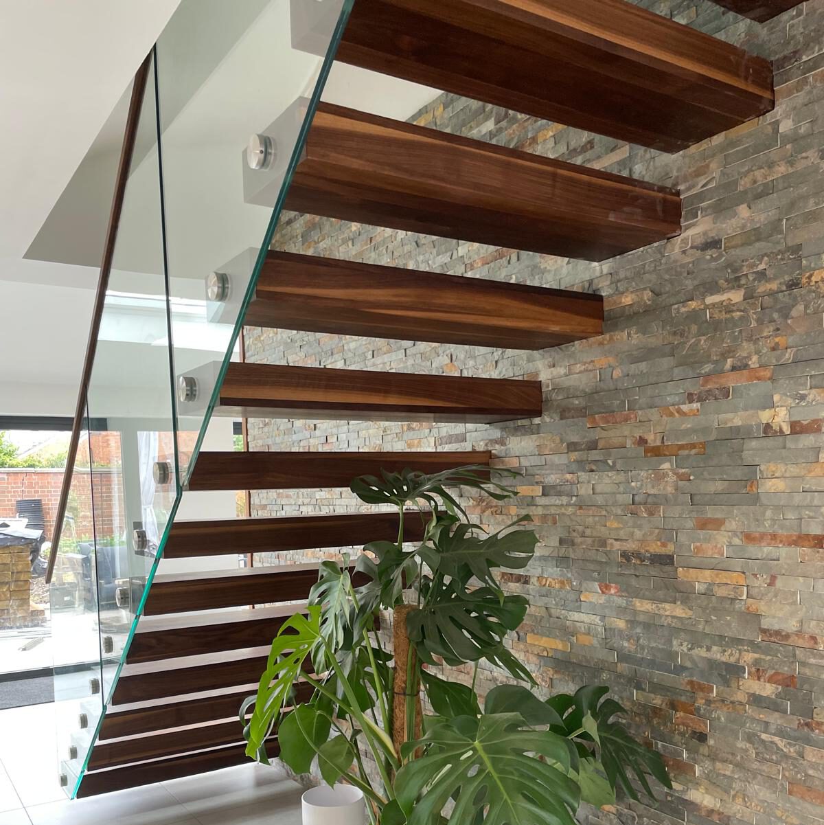Modern floating wooden staircase with glass railing beside a stone-textured wall. Two large potted Monstera plants sit on the white tiled floor underneath the stairs. Natural light fills the space.