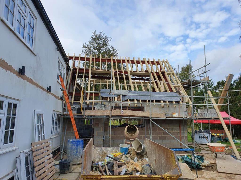 A house under construction with exposed wooden roof beams, scaffolding, and building materials scattered in the foreground. The sky is partly cloudy and trees are visible in the background.