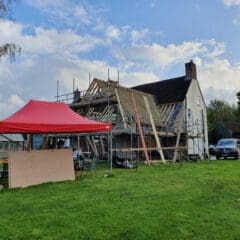 A house under construction with exposed roof beams and scaffolding, a red canopy tent, wooden boards, and tools on the grassy lawn. Two parked cars and a greenhouse are visible in the background under a partly cloudy sky.