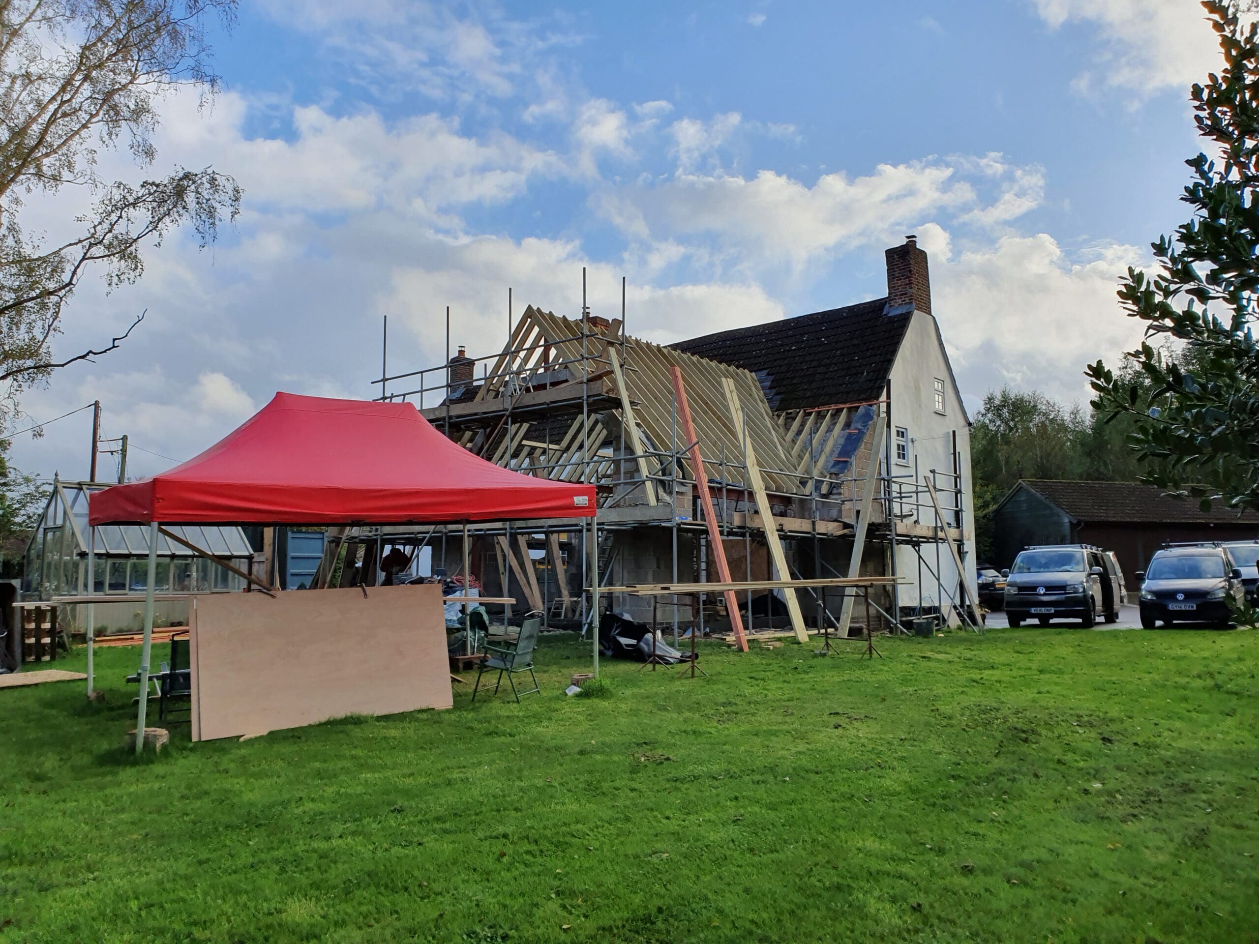 A house under construction with exposed roof beams and scaffolding, a red canopy tent, wooden boards, and tools on the grassy lawn. Two parked cars and a greenhouse are visible in the background under a partly cloudy sky.