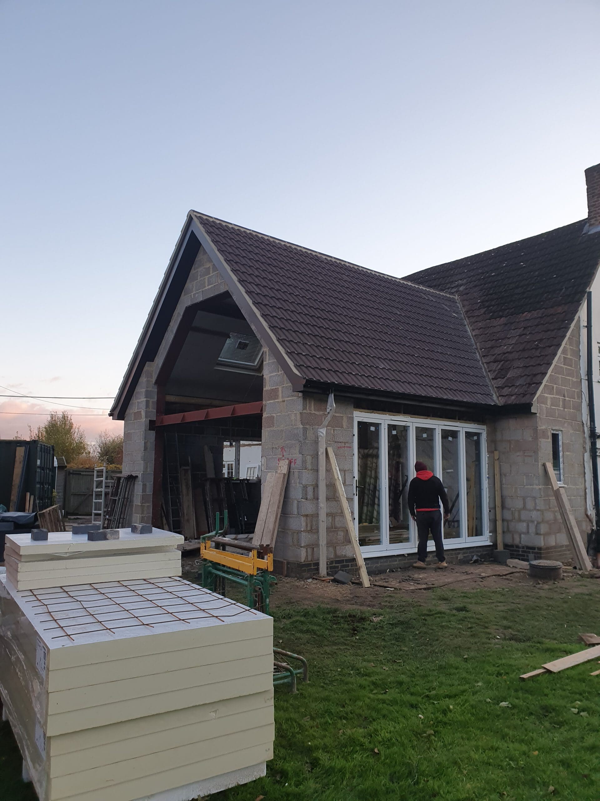 A person stands in front of a partially constructed house extension with large glass doors, unfinished stone walls, and building materials stacked on the lawn.