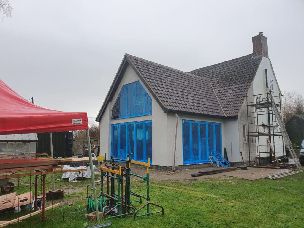 A house under renovation with blue protective coverings on the windows, scaffolding on the right side, and construction materials and a red tent canopy in the yard. The sky is overcast.