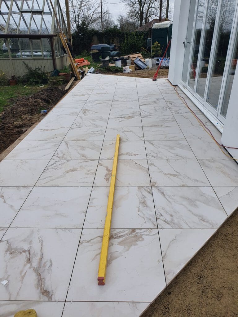 White marble-effect tiles are being installed on an outdoor patio. A long yellow spirit level rests on the tiles. Garden tools, construction materials, and a greenhouse are visible in the background.